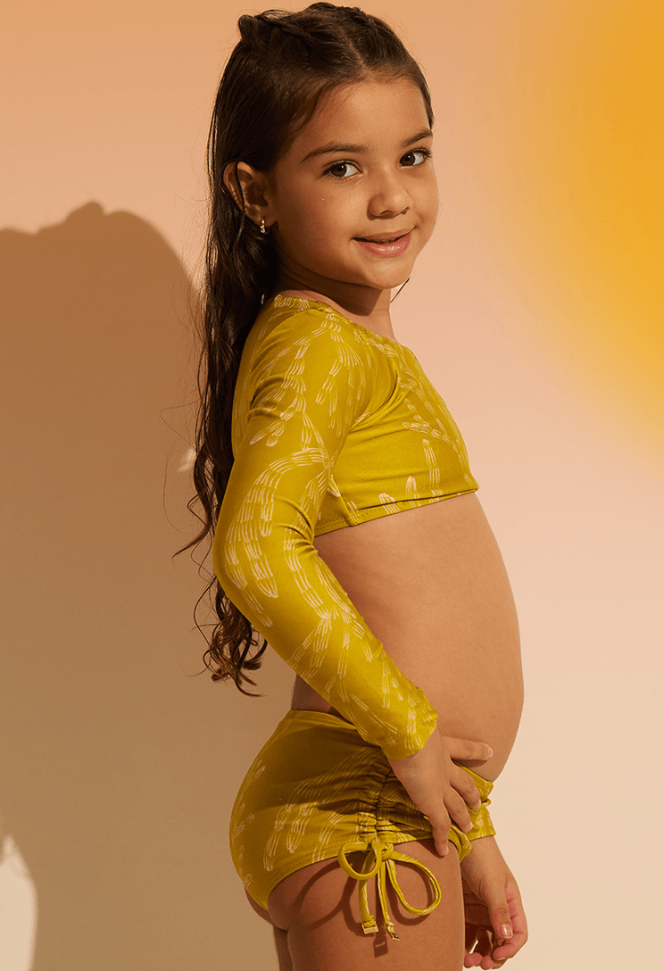 Perfil de menina usando biquíni Valentina Corais amarelo mostarda com estampa branca e blusa de manga longa