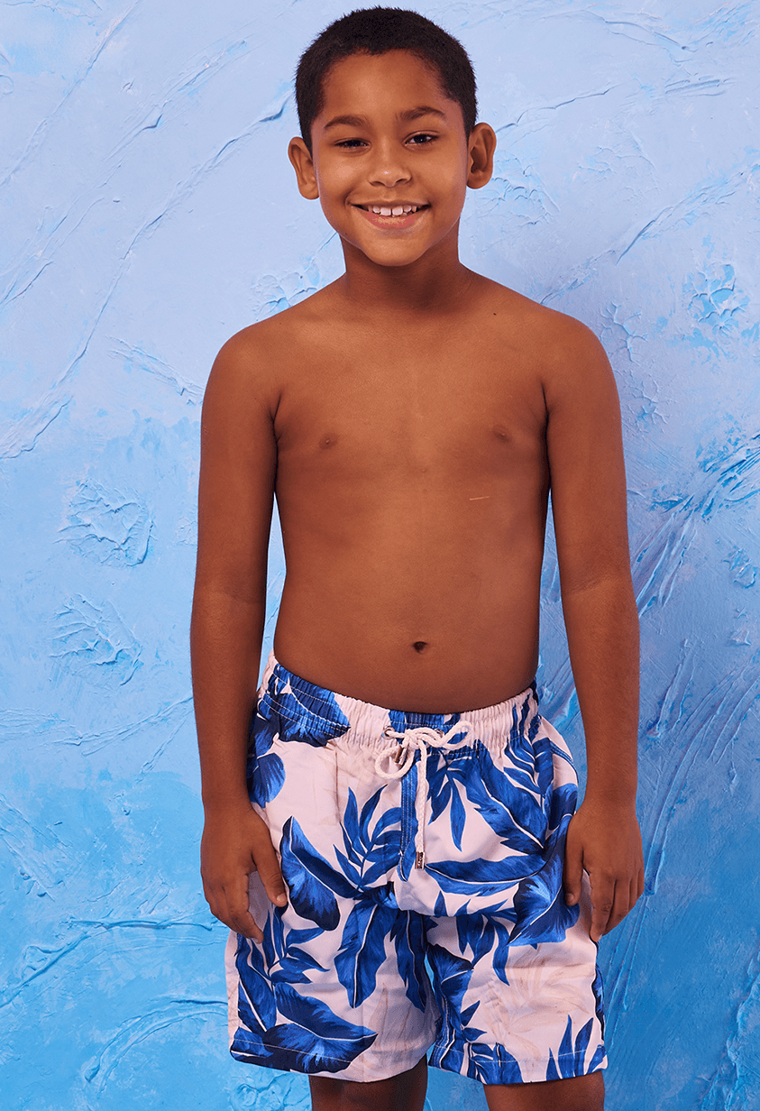 Imagem frontal de menino sorridente vestindo bermuda infantil elástica com estampa de folhas azuis em fundo claro, com cordão branco na cintura