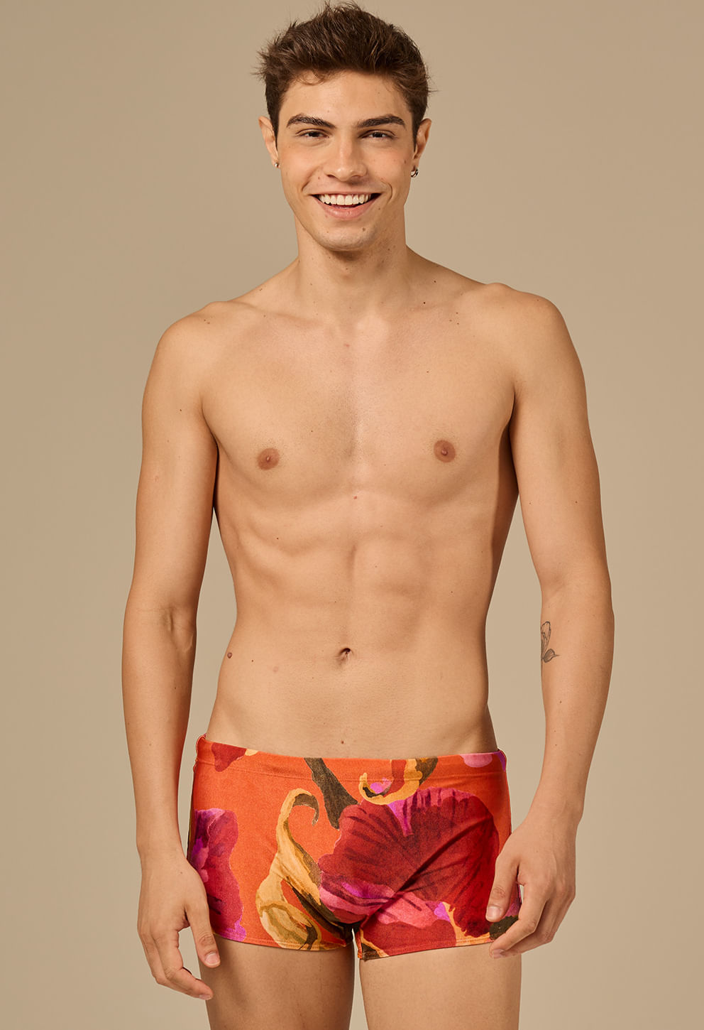 Homem jovem sorridente posando de frente com sunga short adulto chamada 'Chamego'. A sunga tem estampa floral grande com predominância das cores vermelha, laranja, rosa e detalhes em amarelo e verde. 