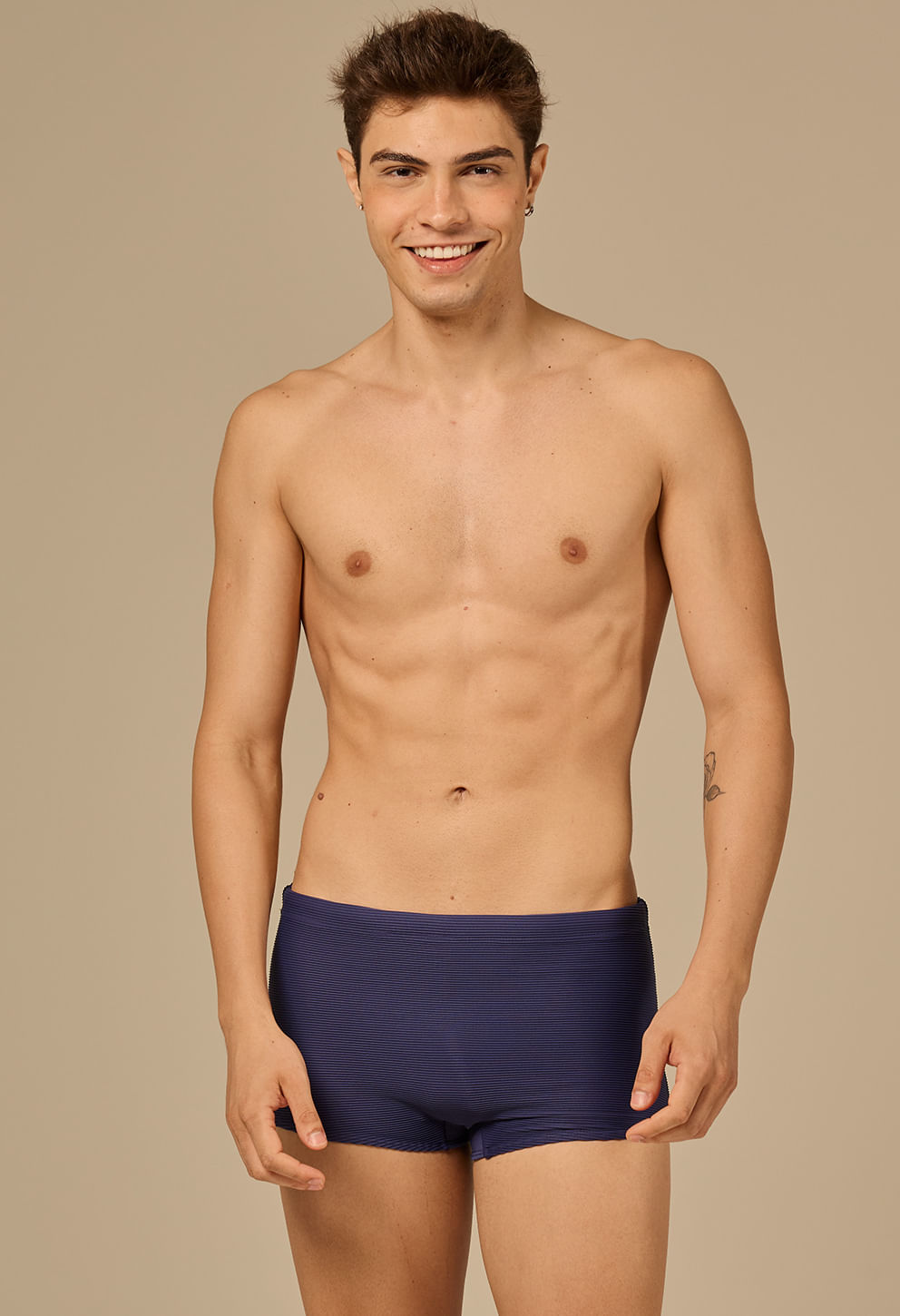 Homem jovem usando sunga short azul marinho com textura listrada, em fundo bege claro