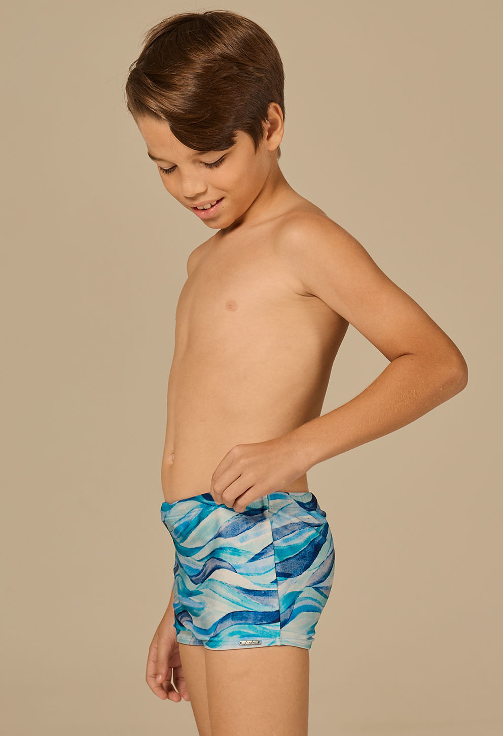 Menino posando de lado usando sunga short infantil estampada em ondas azul claro, azul escuro e branco