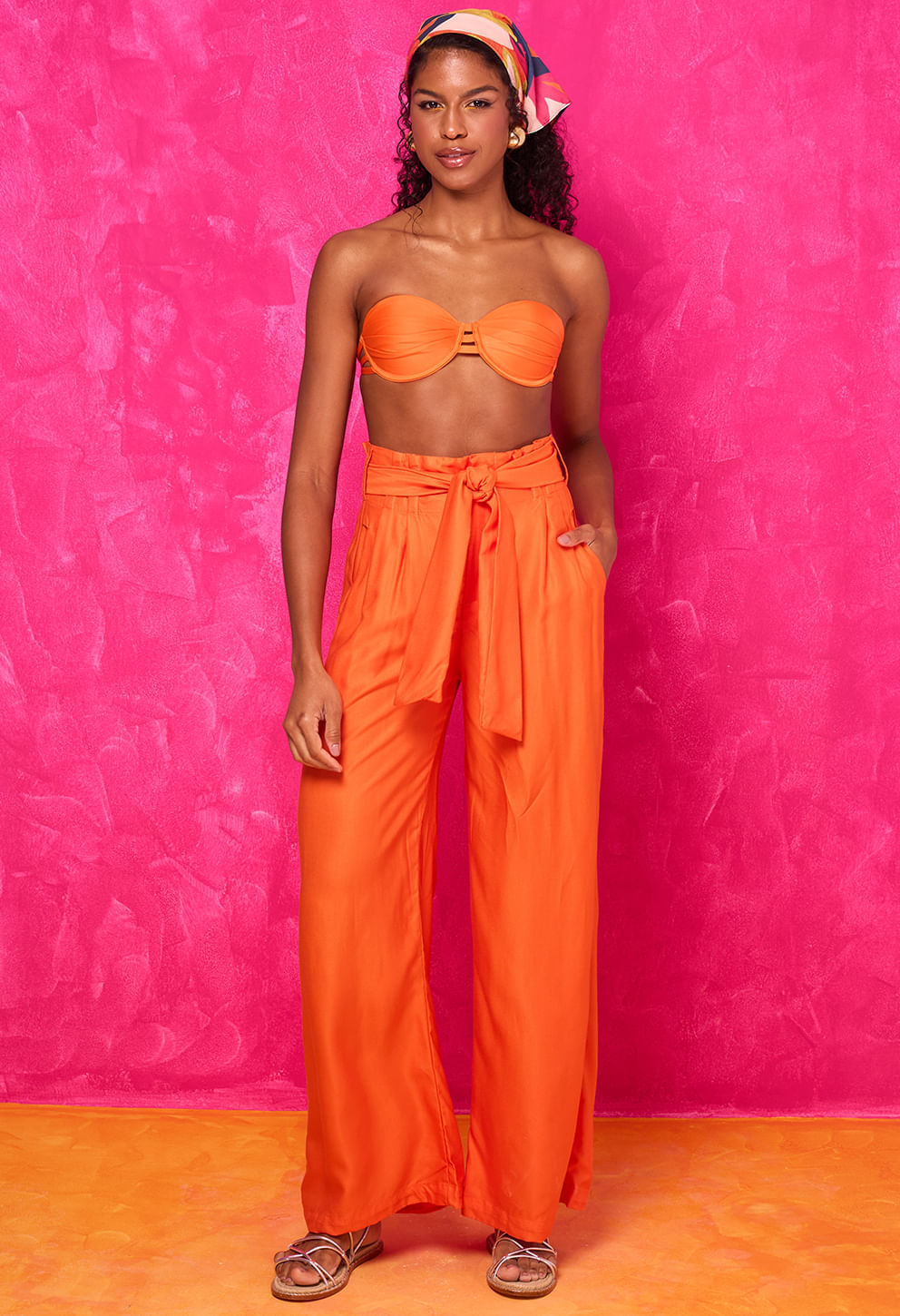 Modelo vestindo Calça Pantalona Glória Bossa laranja vibrante, cintura alta com faixa para amarração e caimento amplo