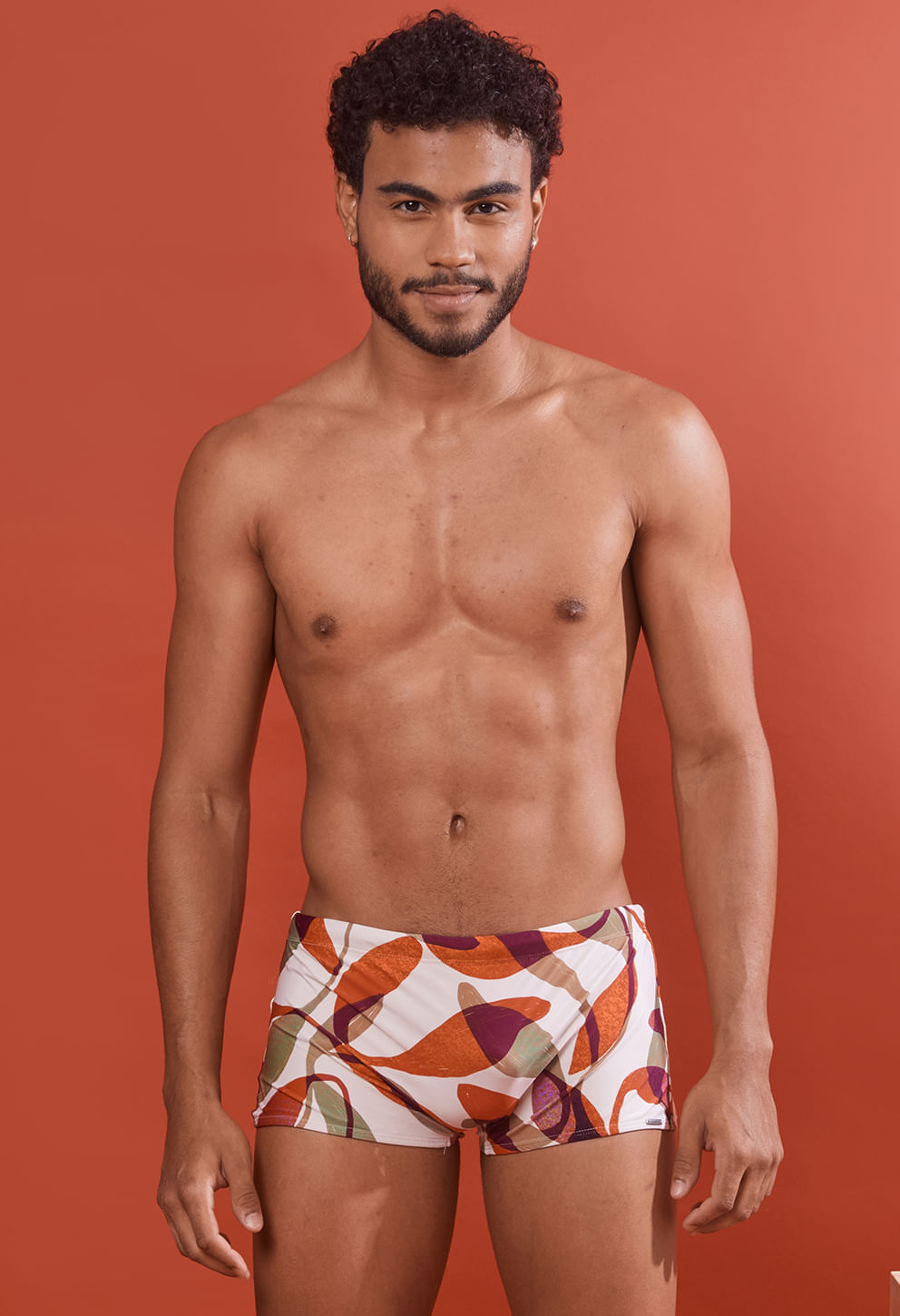 Homem modelo vestindo sunga short adulto com estampa abstrata em branco, laranja, roxo, verde e bege, fundo laranja