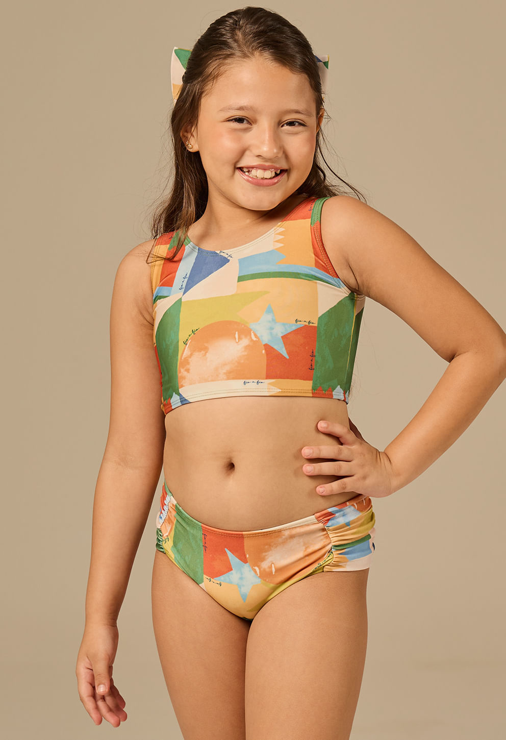 Menina sorridente com cabelo castanho claro preso parcialmente por um laço colorido combinando, vestindo biquíni infantil estampado com padrão geométrico de balões, estrelas e cores vivas como laranja