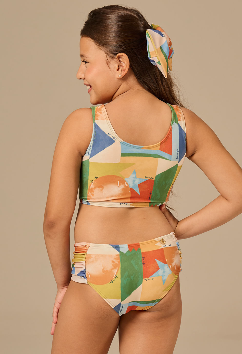 Menina de perfil e costas para a câmera, vestindo biquíni infantil estampado colorido com balões e estrelas em tons de laranja, verde, azul e amarelo