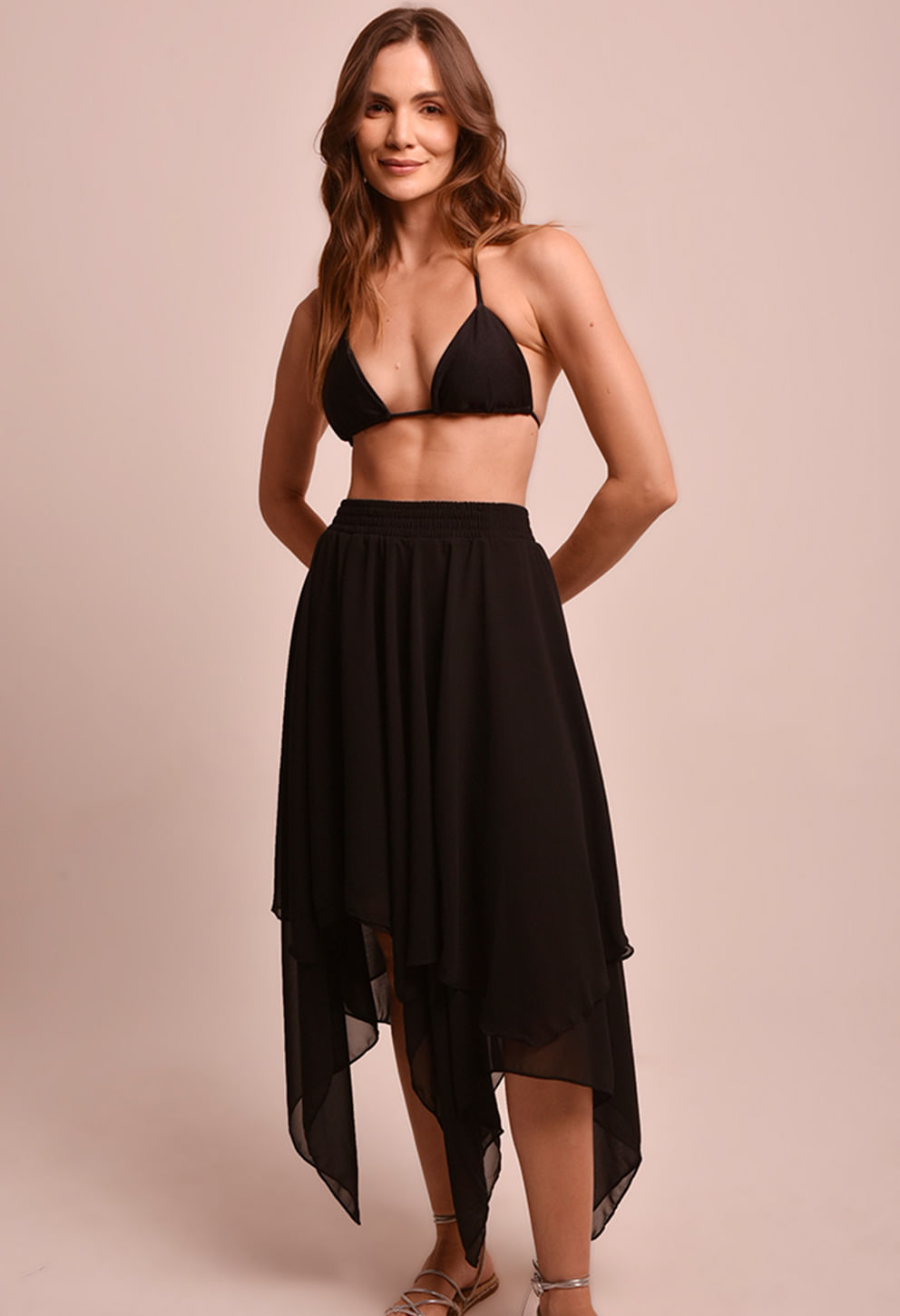 Modelo feminina em estúdio usando saia preta assimétrica com camadas de tecido leve e cós elástico. Visual frontal com top de biquíni preto; fundo rosa claro neutro.