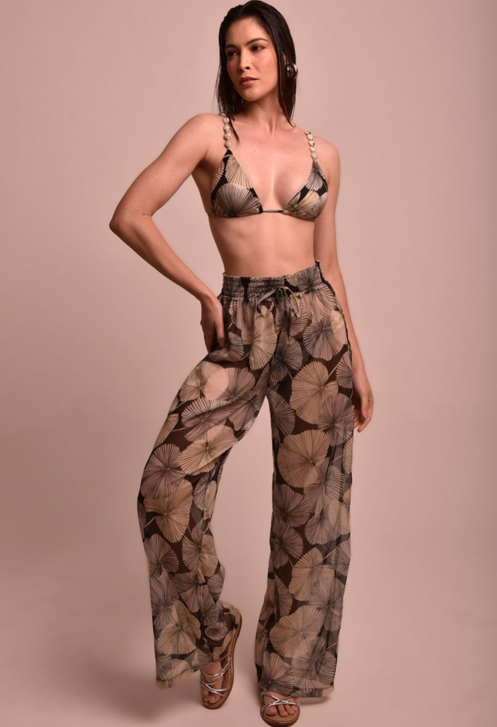 Modelo em pé usando calça pantalona estampada em tons bege, marrom e preto com padrão de folhas/abanicos; cintura alta elástica com cordão; tecido leve, semitransparente, caimento fluido e perna larga