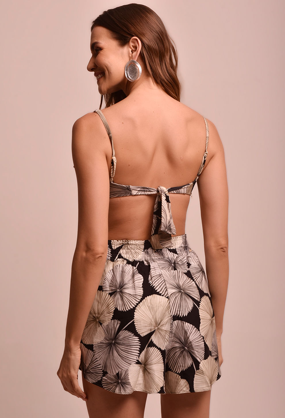 Mulher jovem vista de costas contra fundo bege claro, usando top de alças finas e short com estampa de folhas em tons neutros
