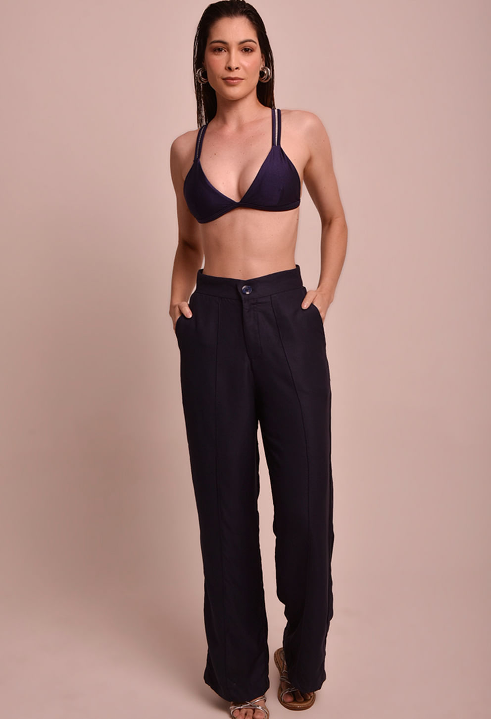 Imagem frontal de uma modelo usando uma calça pantalona na cor marinho, cintura alta com botão e zíper, bolsos laterais e costuras verticais que alongam a silhueta.