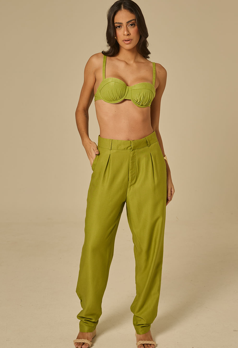 Modelo feminina em pé frente, usando calça reta verde esmeral de cintura alta com pregas frontais, passantes, bolsos laterais e dobra discreta na barra.