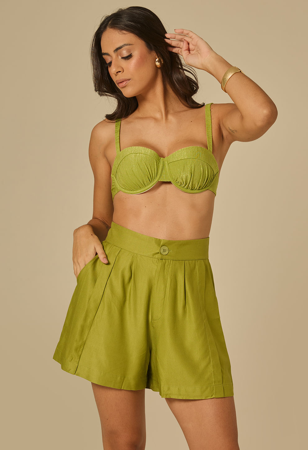 Modelo feminina em ângulo frontal usando short Milão na cor esmeral (verde), cós alto com botão frontal, pregas frontais e bolsos laterais.