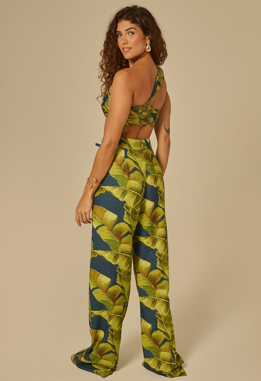 Vista traseira da calça pantalona estampada com folhas tropicais verdes, caimento amplo e fechamento no cós, em fundo bege neutro.