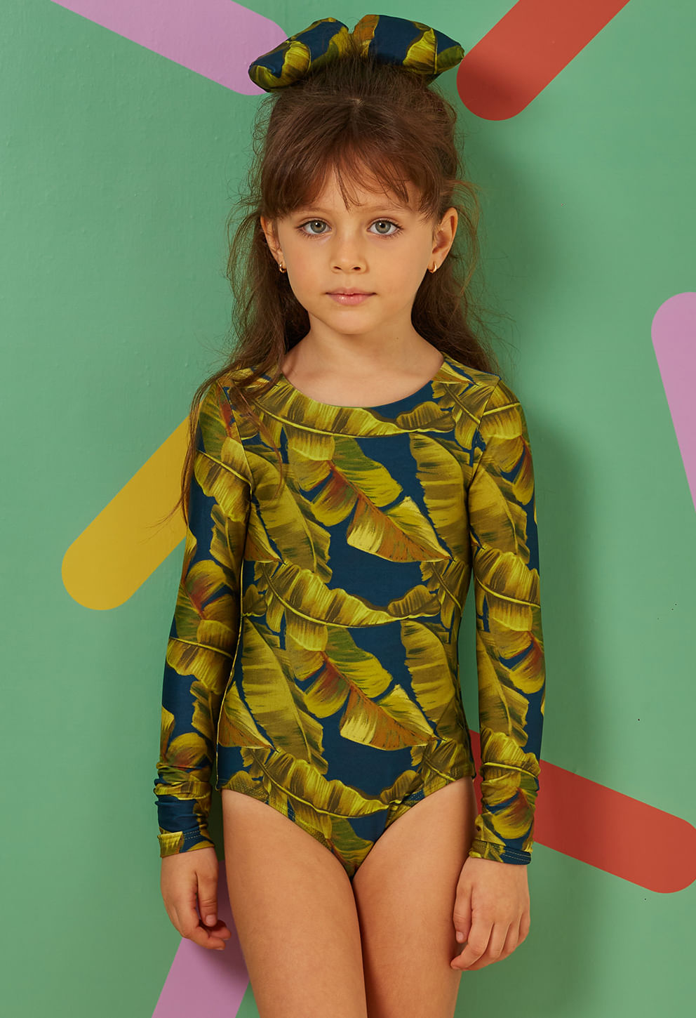 Menina infantil usando maiô de manga longa com estampa de folhas grandes em tons verde-oliva e fundo azul petróleo
