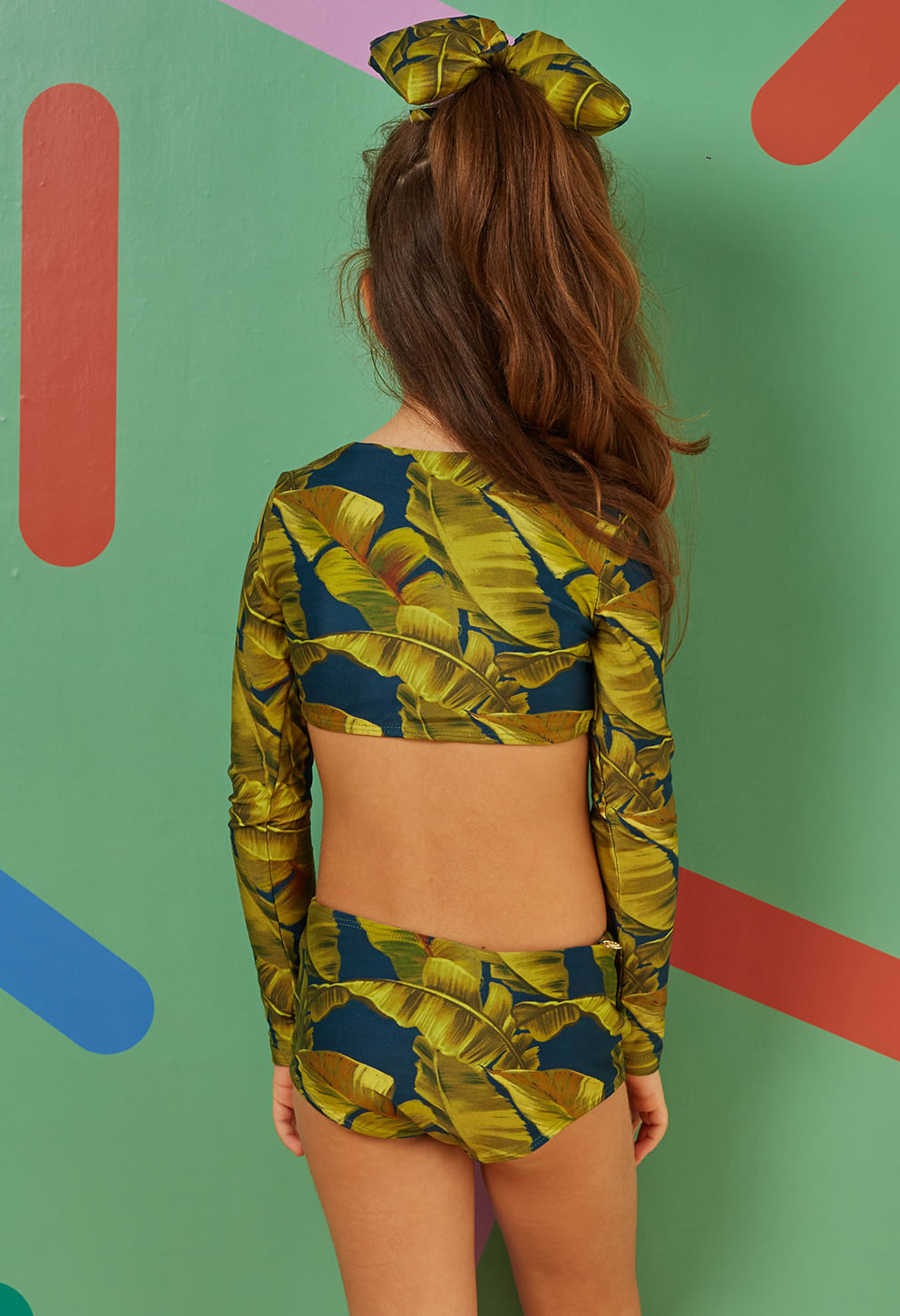 Vista traseira de menina usando biquíni infantil de mangas longas e hot pants com estampa tropical de folhas em verde e amarelo sobre base azul escuro.
