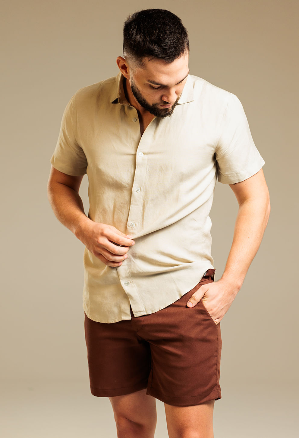 Modelo masculino em foto frontal usando bermuda alfaiataria em tom marrom 'Godiva', com camisa clara manga curta.