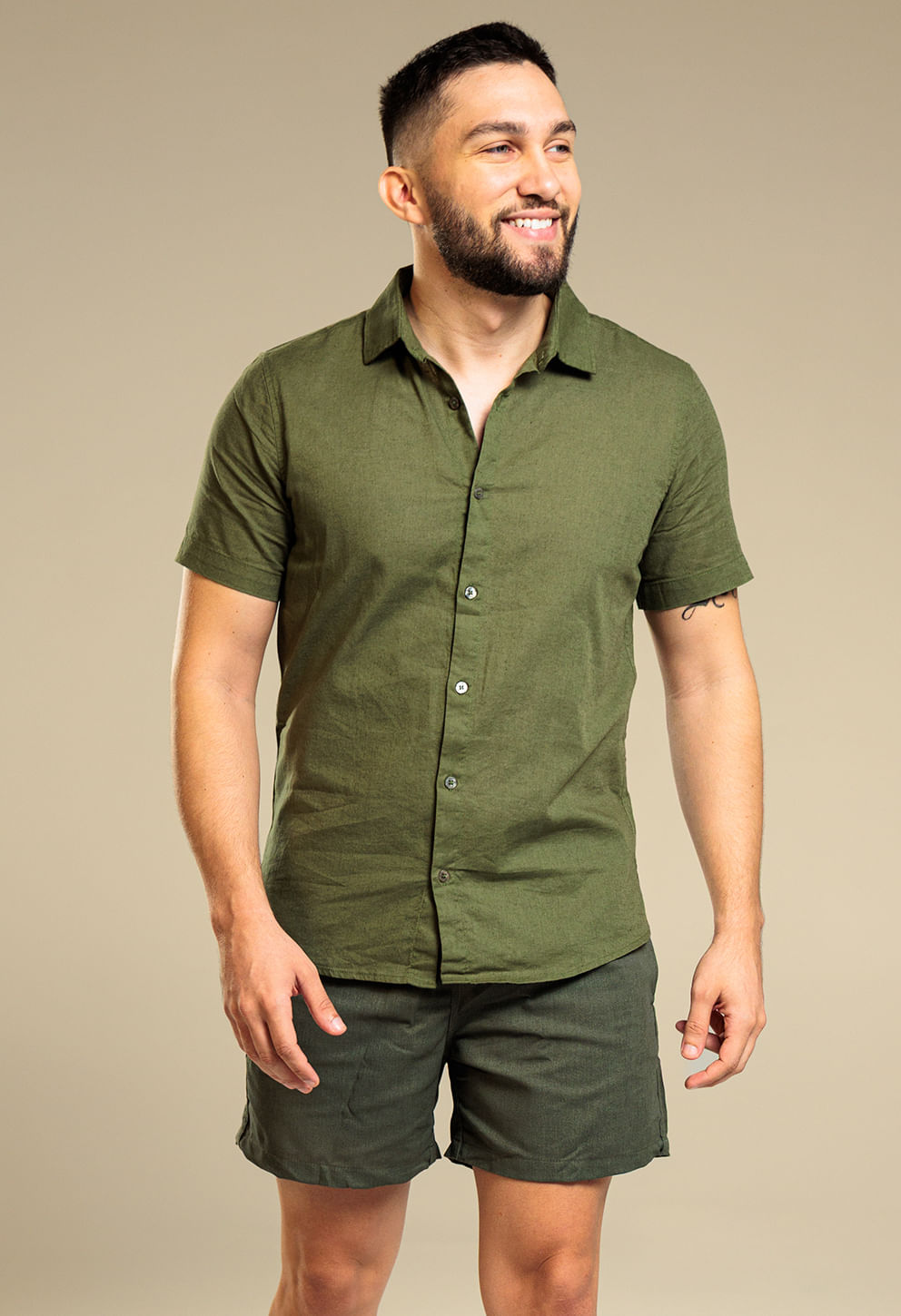Modelo masculino em pé, três-quartos frontal, veste camisa de manga curta verde oliva e bermuda alfaiataria verde escuro