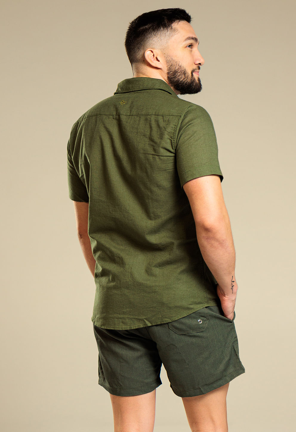 Visão traseira de modelo masculino usando camisa e bermuda verdes; bermuda mostra bolso traseiro com aba e fivela de ajuste lateral