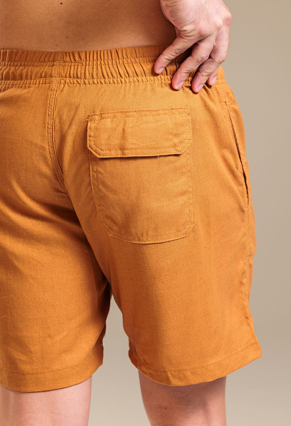 Close nas costas da bermuda camel mostrando cós elástico, costura reforçada e bolso traseiro quadrado com aba; tecido com textura semelhante a linho e caimento descontraído; mão do modelo ajustando a