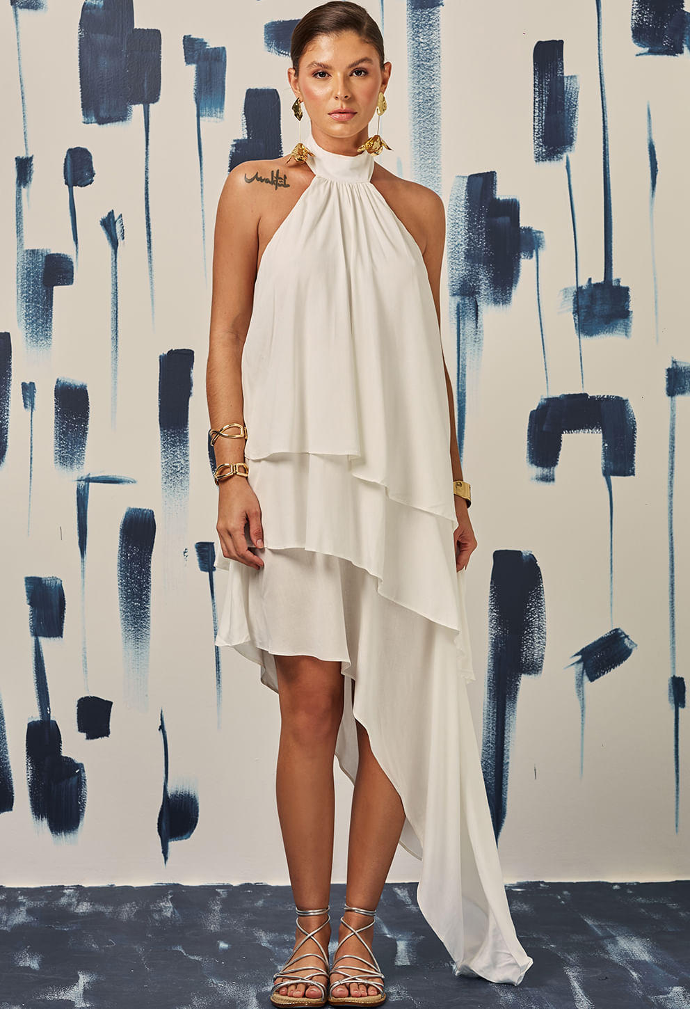 Modelo feminina usando vestido off white halter com sobreposições em camadas assimétricas, bainha longa lateral, costas nuas e decote fechado no pescoço