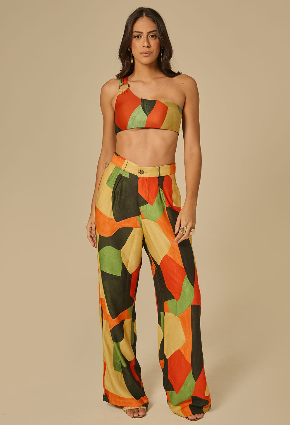 Modelo feminina em pé com calça pantalona social estampada geométrica predominante em tons mostarda, laranja, verde e preto.