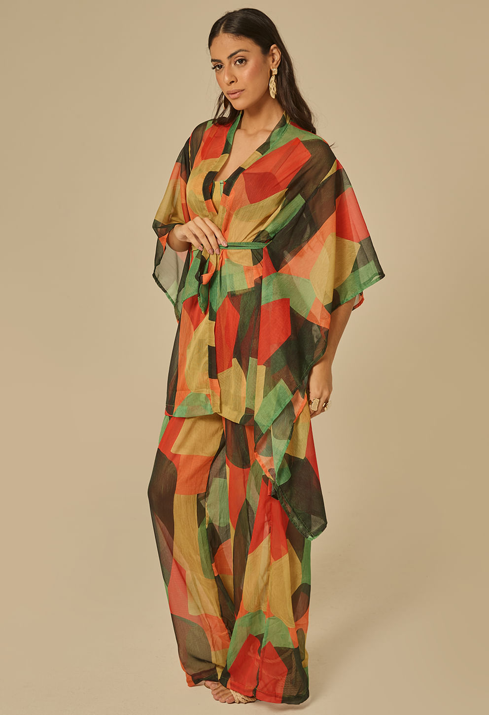 Modelo feminina em kimono longo translúcido com estampa geométrica multicolor (verde, vermelho, mostarda e preto). Corte oversized com mangas amplas e cinto fino na cintura; tecido leve e fluido.
