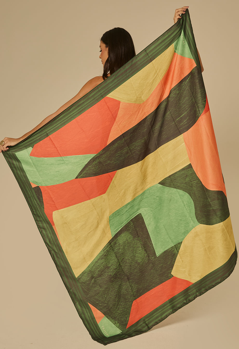 Pareô longo com estampa geométrica em blocos verdes, laranja, amarelo e detalhes em preto; tecido leve e translúcido, amarrado e sustentado por modelo que segura a peça estendida.