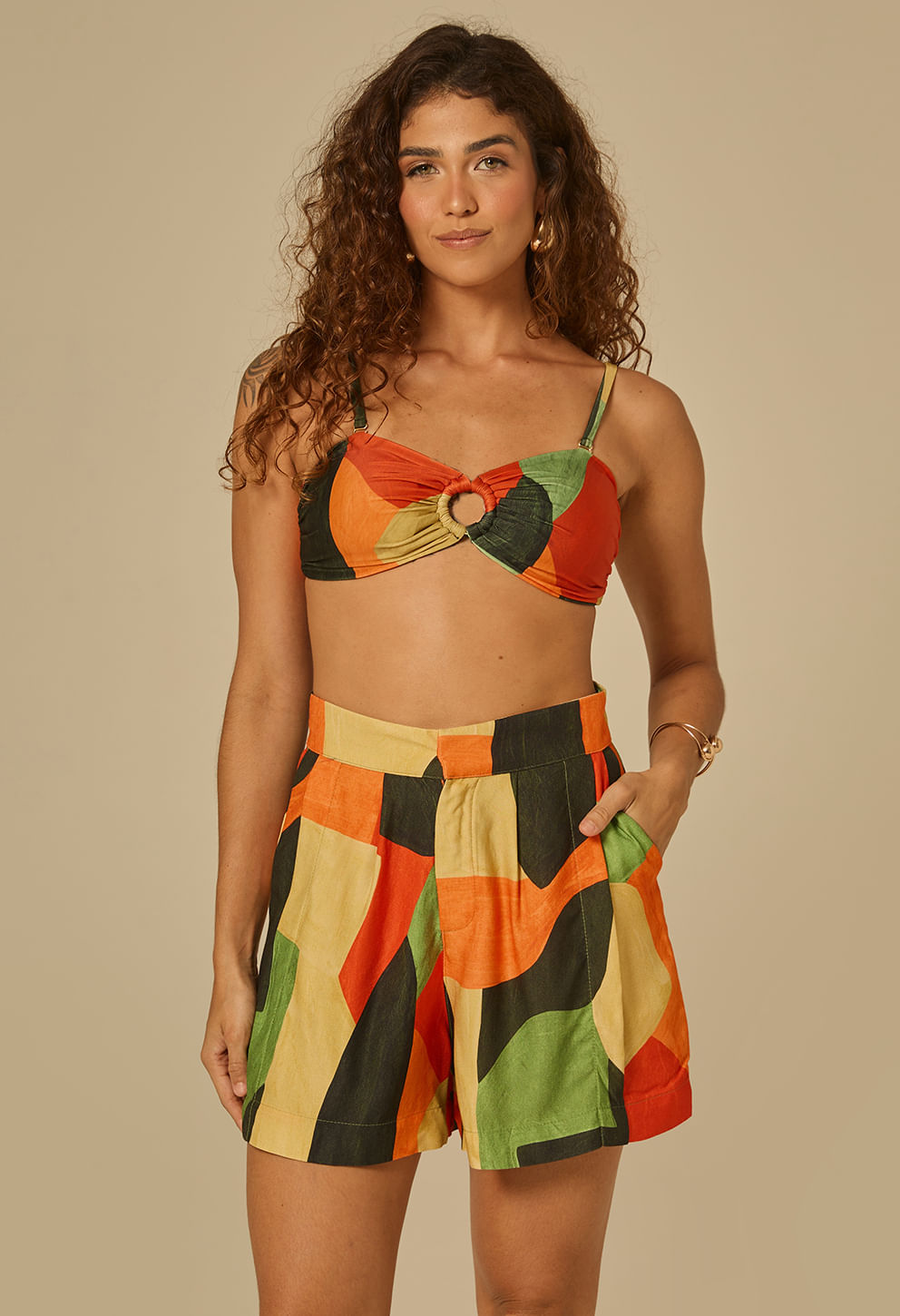 Modelo feminina de frente usando short curto estampado geométrico em tons predominantes verde, laranja, preto e bege