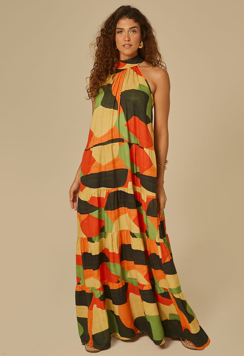 Vestido longo feminino estampado em tons predominantes de laranja, verde, preto e bege. Decote halter alto com franzido no peito, caída ampla em camadas com leve godê.