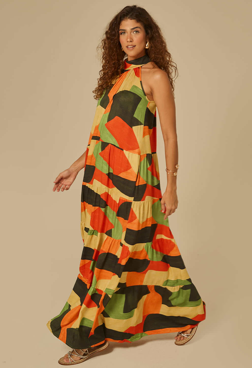 Vestido longo feminino em movimento, estampa geométrica multicolor (laranja, verde, preto, bege). Decote halter com gola alta levemente drapeada.