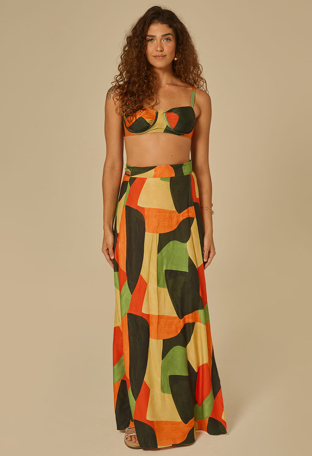 Modelo de frente usando saia longa transpassada com estampa geométrica abstrata em tons verde, laranja, preto e amarelo.