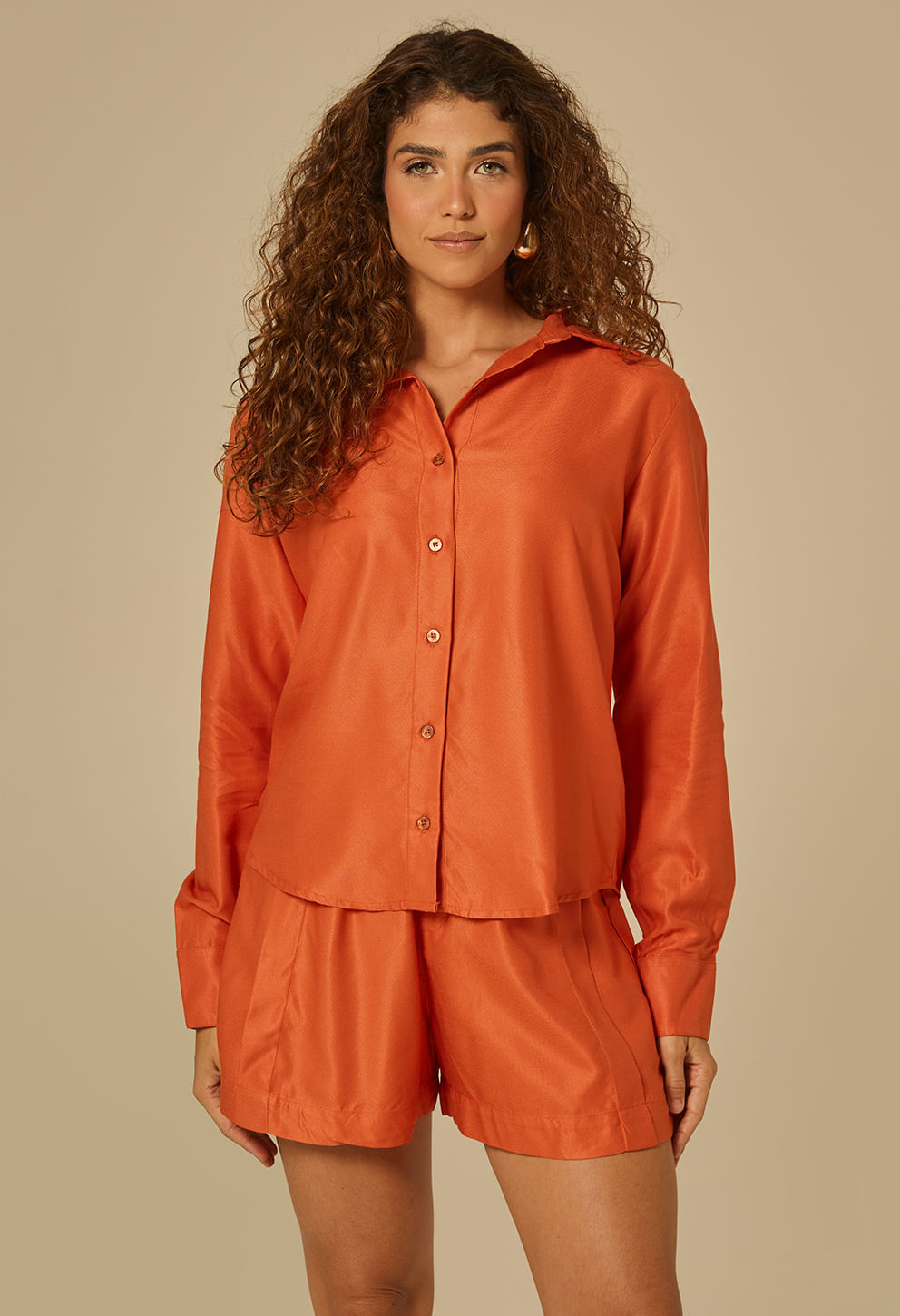 Modelo feminina em fundo neutro usando camisa botão laranja queimado com caimento fluido e brilho acetinado