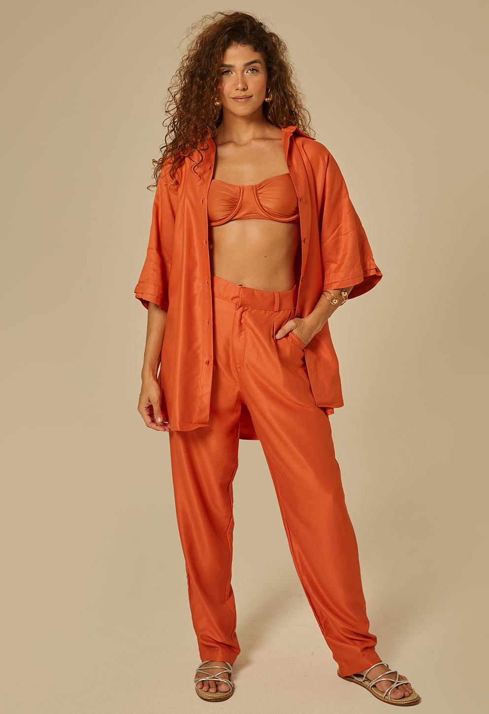 Modelo com camisão aberto mostrando top de biquíni por baixo e calça com elástico; look praia/resort em tom laranja queimado acetinado