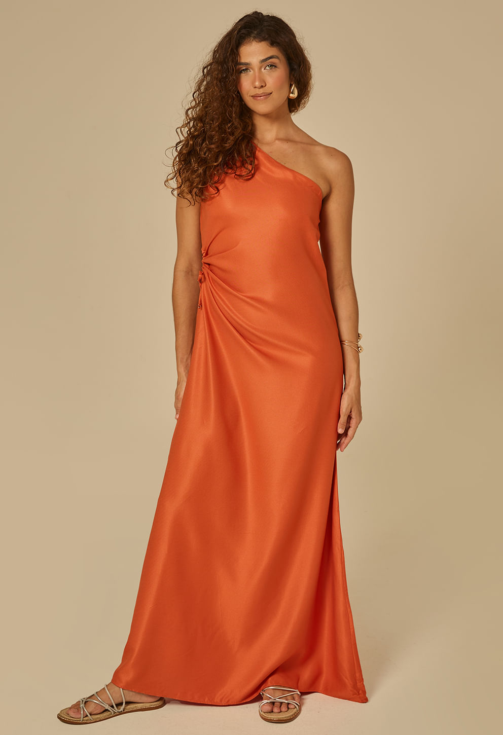 Modelo feminina em estúdio neutro vestindo vestido longo laranja tom sunset, um ombro só com franzido lateral ajustável e corte levemente fluido.