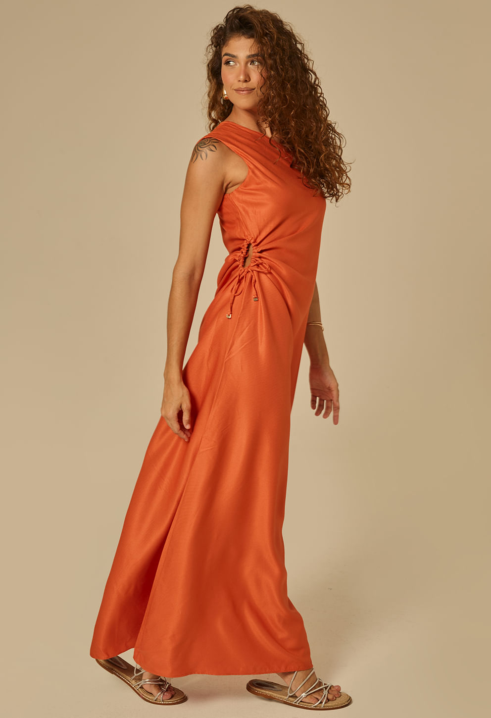 Imagem lateral da modelo mostrando detalhe do franzido lateral ajustável tipo drapeado com cordões; vestido longo laranja, ombro único estilizado, caimento fluído e acabamento acetinado.