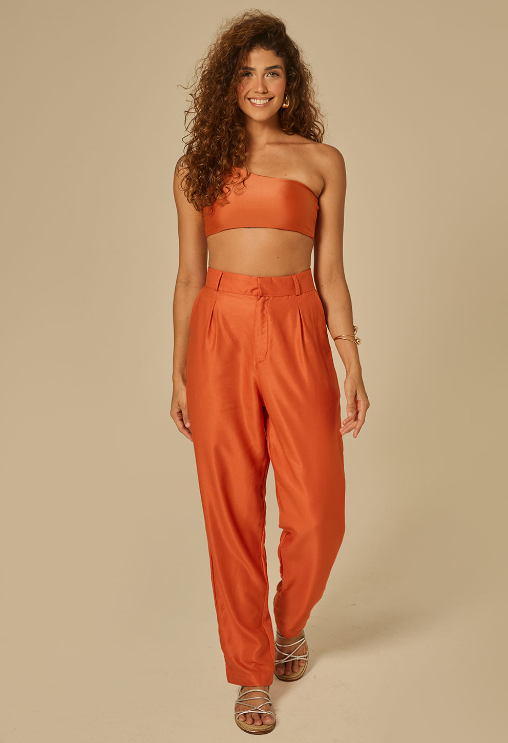 Modelo feminina usando calça reta laranja queimado com cintura alta e pregas frontais, tecido com leve brilho.
