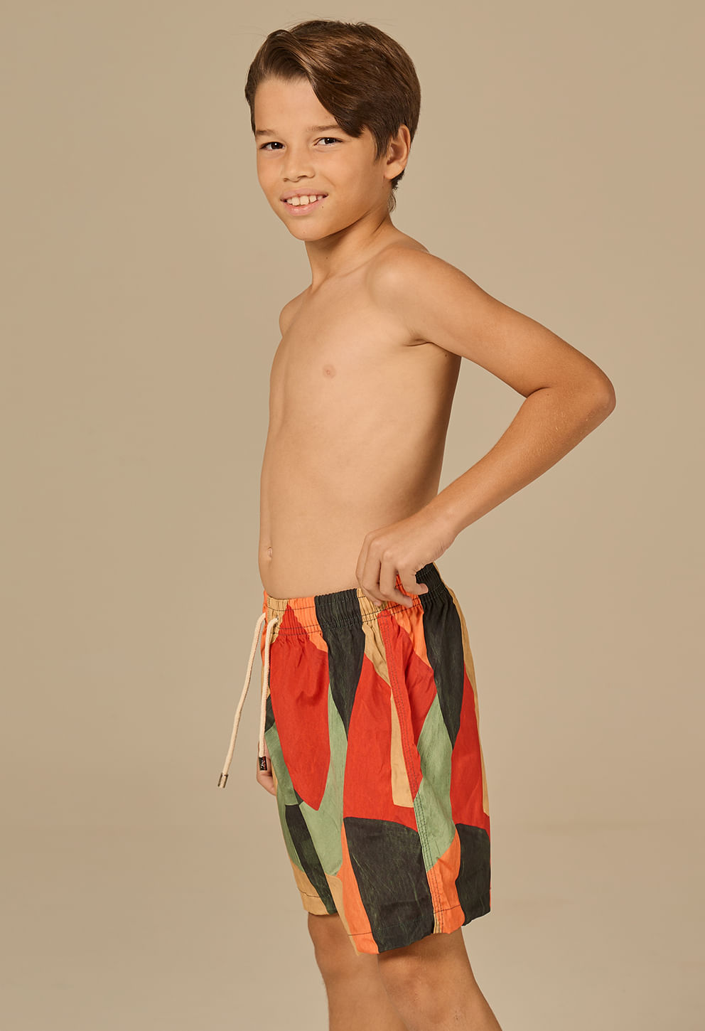 Menino infantil de perfil direito, sem camisa, usando bermuda beach estampada em blocos laranja, vermelho, verde, preto e bege. Cós elástico com cordão longo branco, bolso lateral discreto.