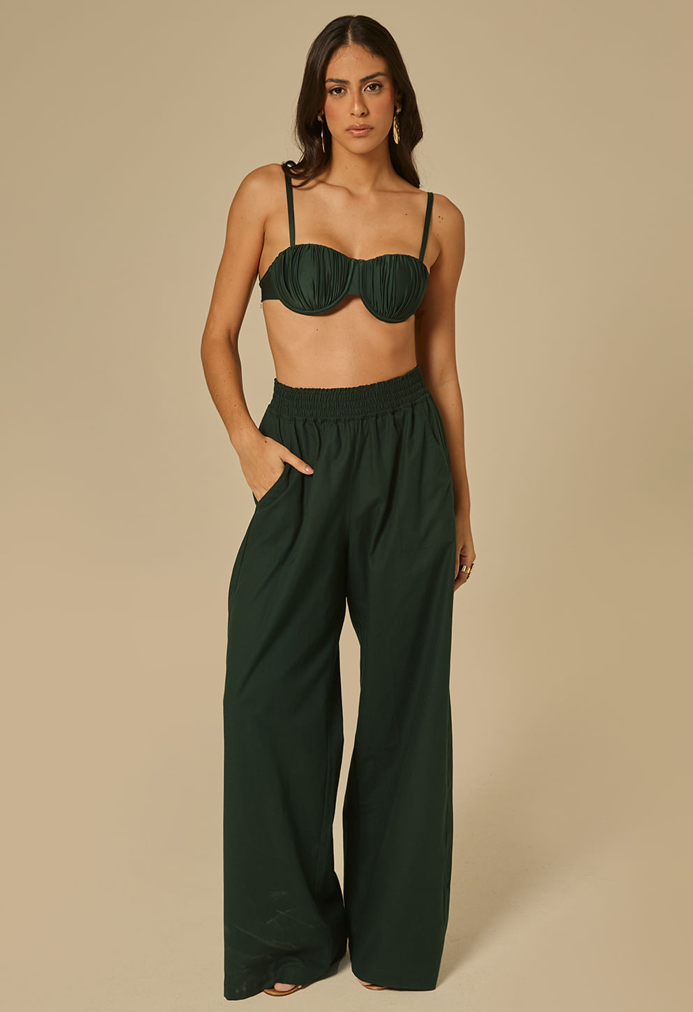 Modelo feminina usando calça verde escuro de cós alto com elástico franzido e bolsos laterais, caimento amplo e perna reta.