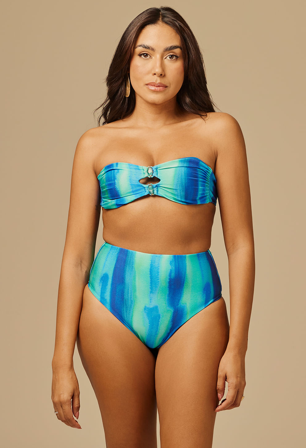 Mulher morena usando biquíni tomara que caia com calcinha hot pants azul e verde com argolas, fundo neutro bege