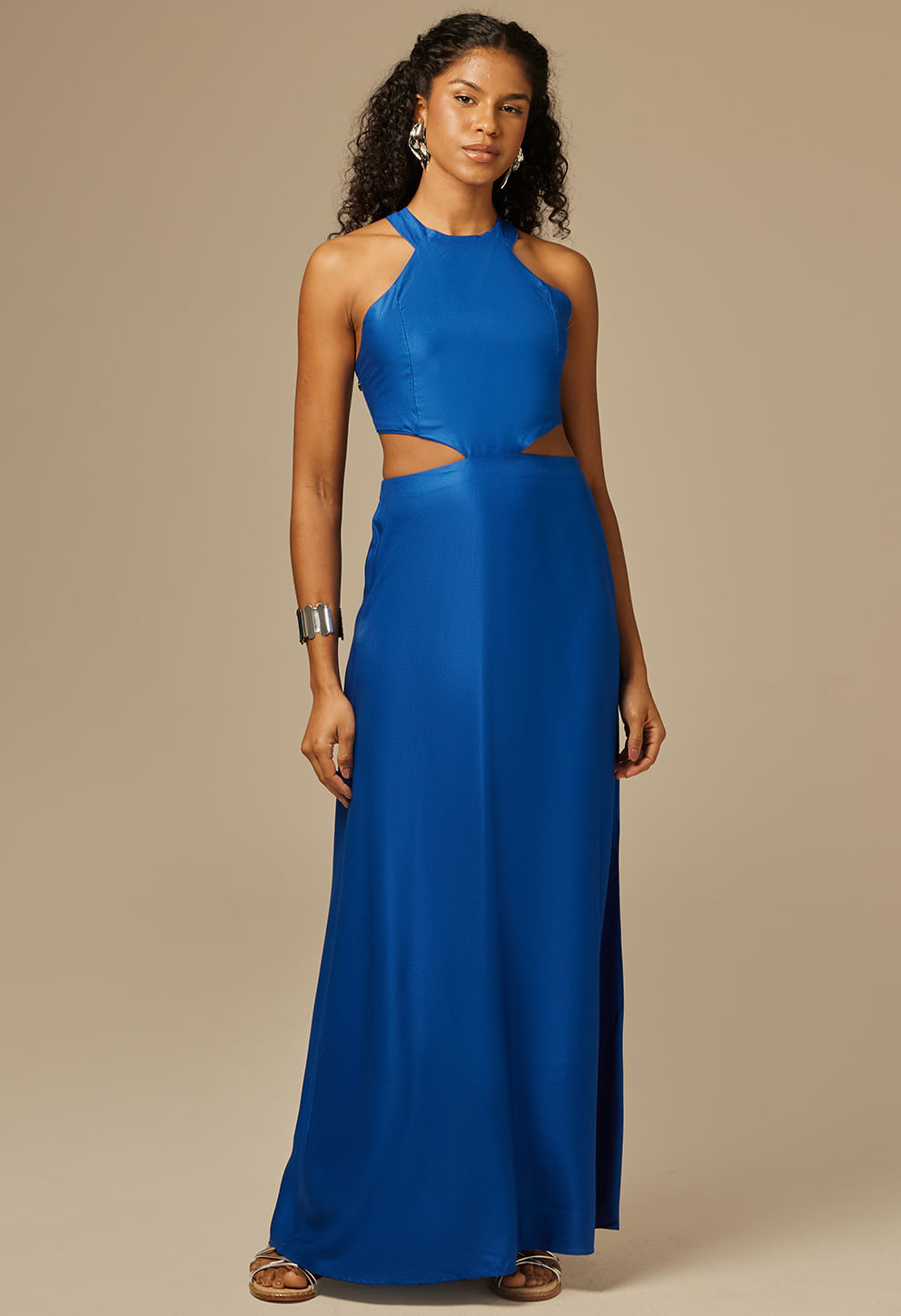 Vestido longo feminino azul royal com modelagem evasê, sem mangas, gola alta e recortes laterais na cintura. O vestido tem caimento fluido e tecido liso, com detalhe de amarração nas costas.