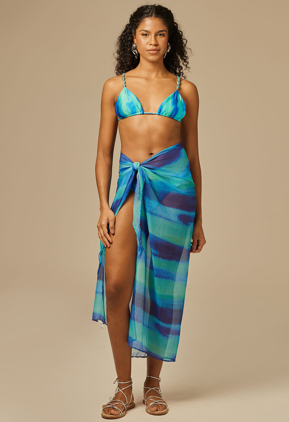 Modelo feminina usando um pareô longo translúcido com estampa em tons de azul, verde e roxo, amarrado na frente com nó